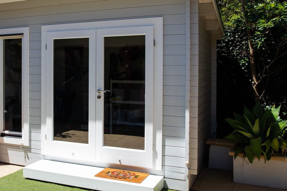 Light grey shed with white-framed French glass doors, a small wooden step, and a doormat in a sunny garden setting. — Daniher Building Company in Belconnen, ACT