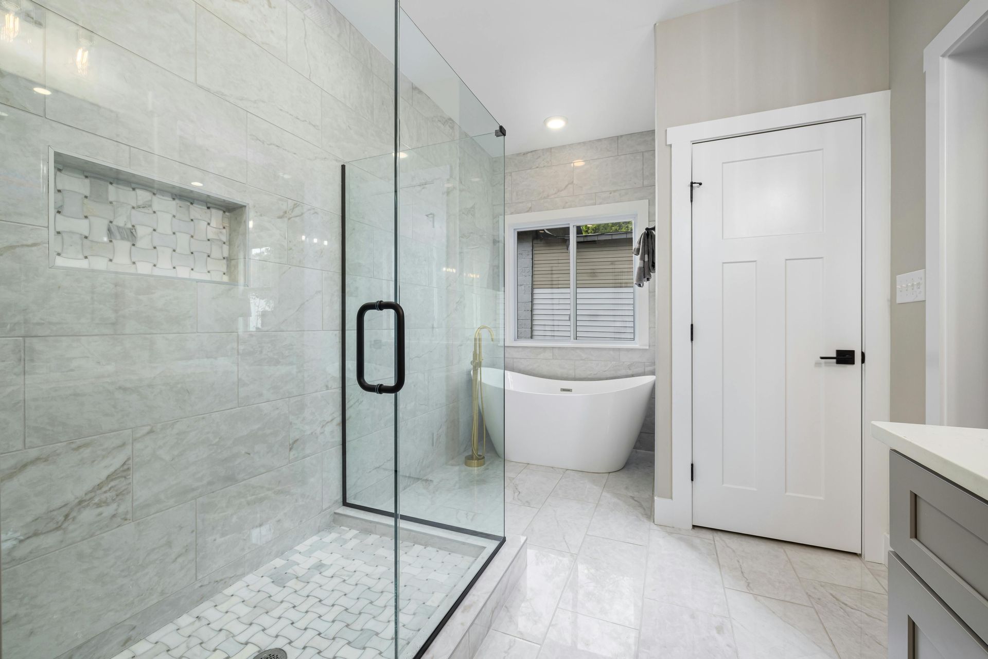 A modern bathroom with a glass-enclosed shower, a white soaking tub by a window, and a white paneled door. — Daniher Building Company in Yass, NSW