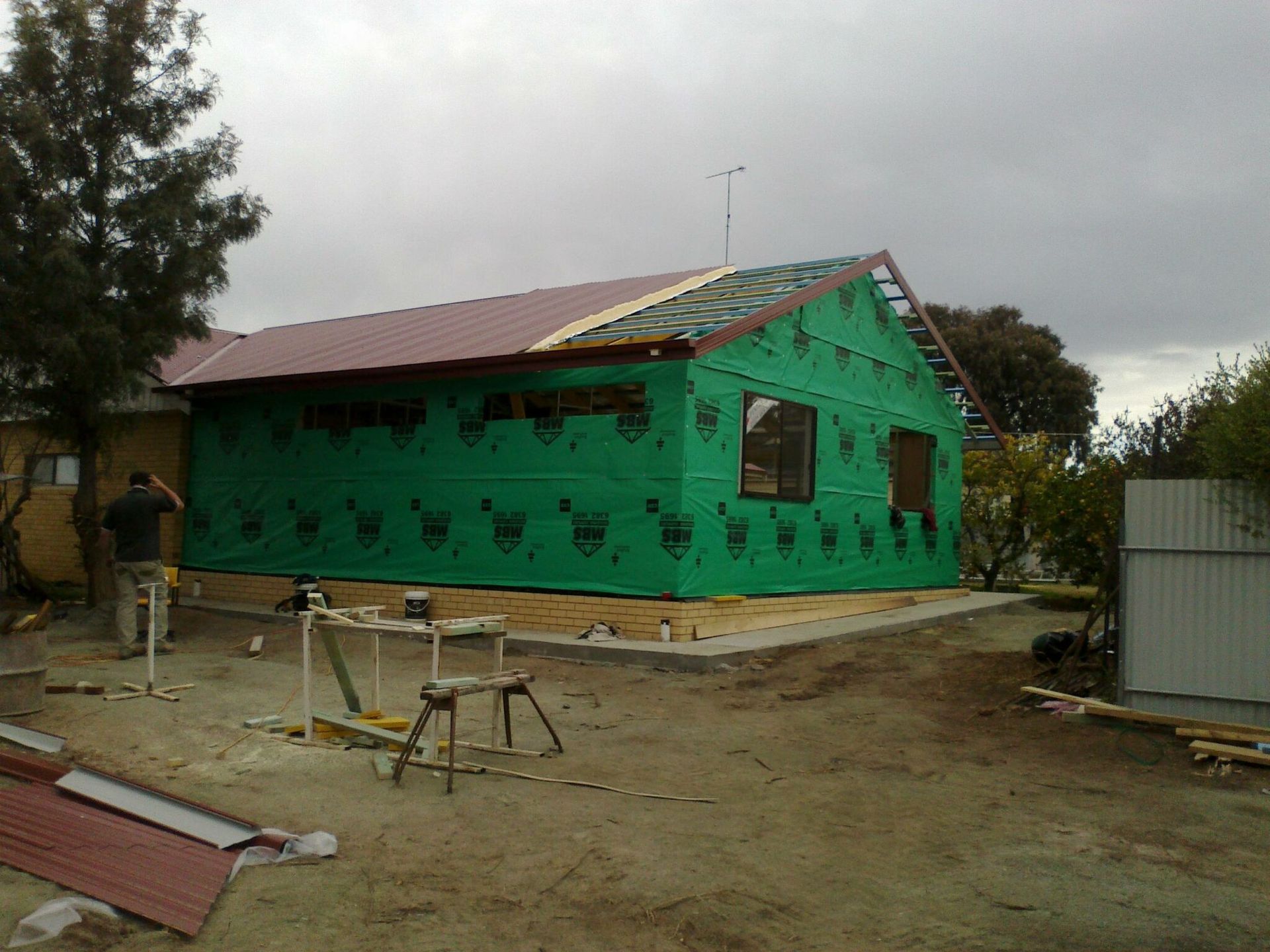 A person stands near a house under renovation, wrapped in green protective exterior sheeting with a partially red roof. — Daniher Building Company in Flynn, ACT