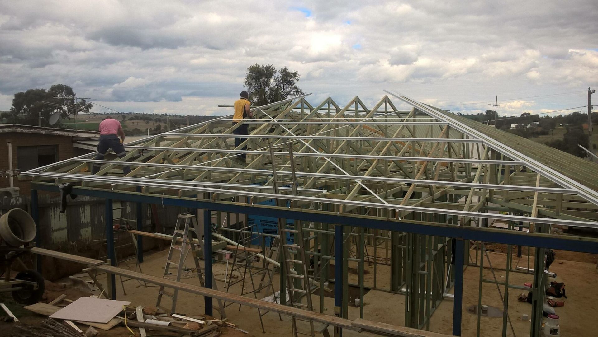 Two workers assemble a metal roof frame on the walls of a house under construction under a cloudy sky. — Daniher Building Company in Flynn, ACT