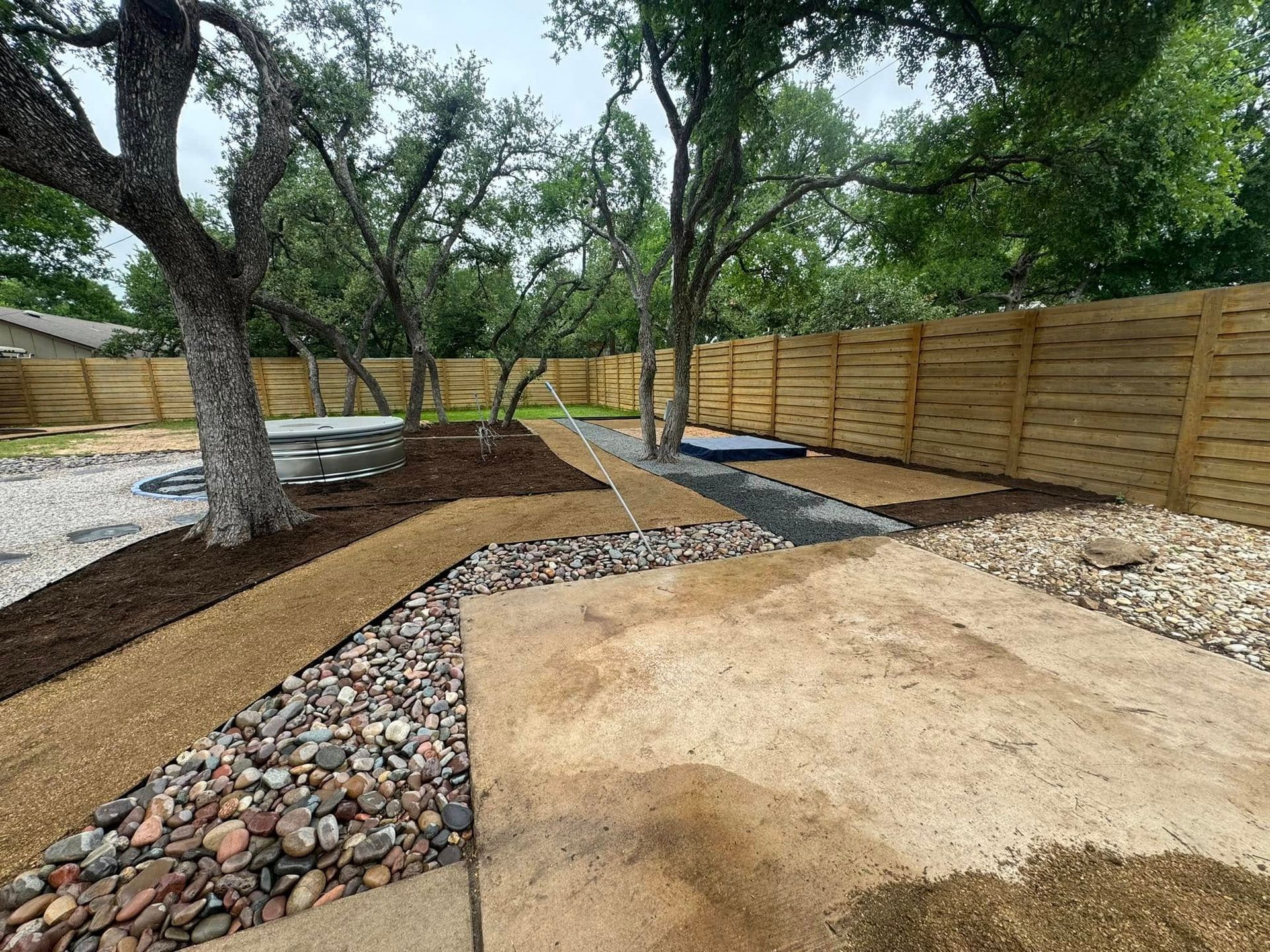 A backyard with a wooden fence , trees and rocks.