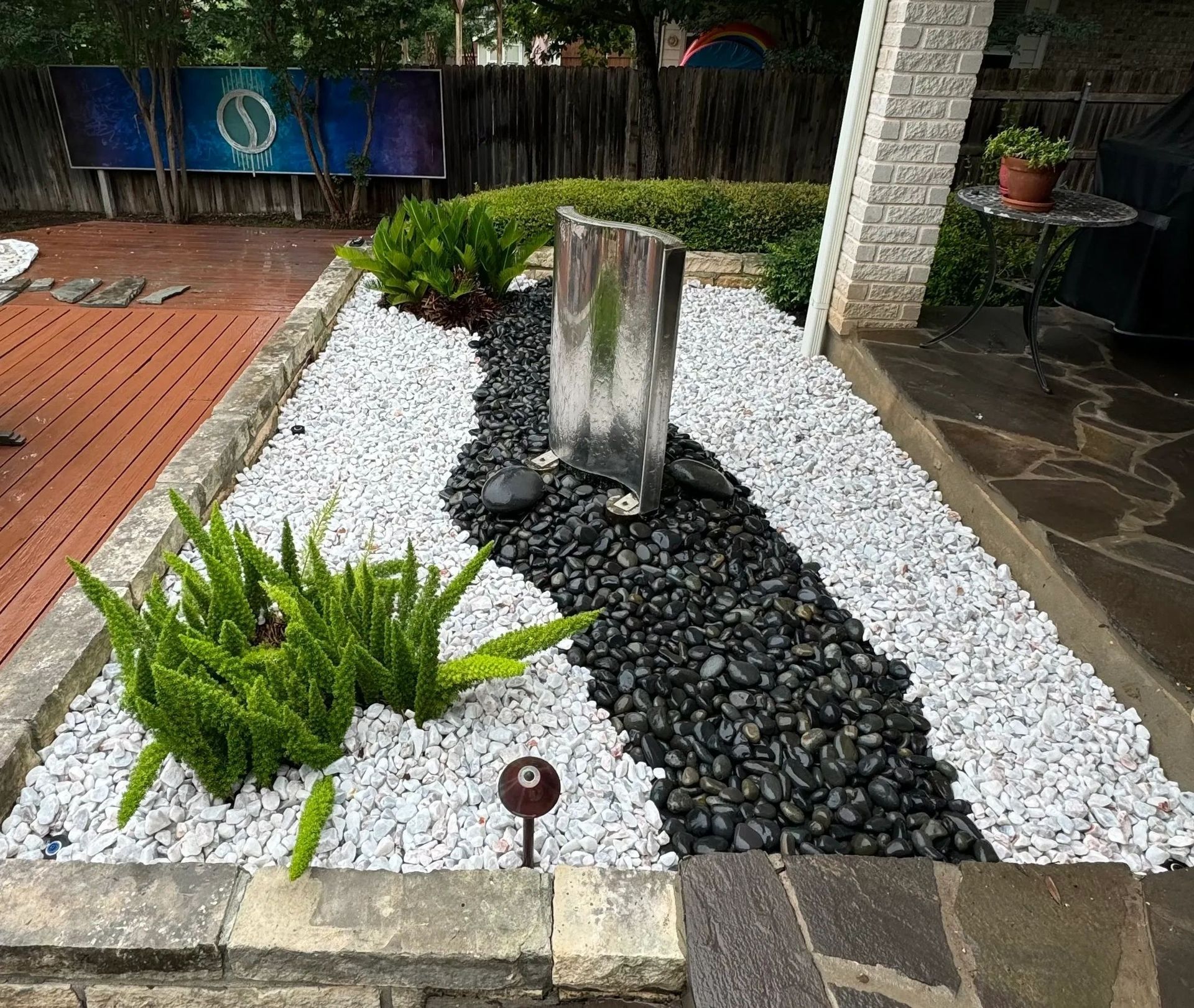 Landscaped garden bed with black and white rocks, water feature, and greenery.