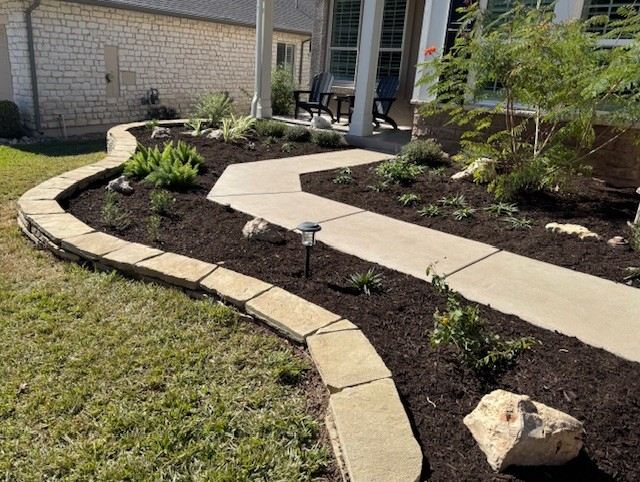 Front yard landscaping with a stone border, mulch, plants, and a concrete walkway.