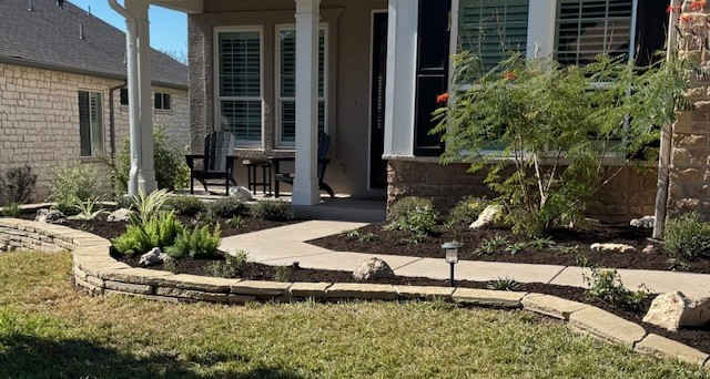 Stone-bordered flowerbeds with plants lead to a home's porch with seating. A walkway curves through the grass.