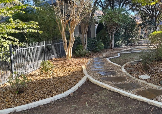 Stone path winds through landscaped yard with a black fence, trees, and a house in the background.