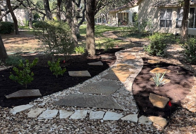 Stone pathway winding through a garden with dark mulch, trees, and small shrubs.