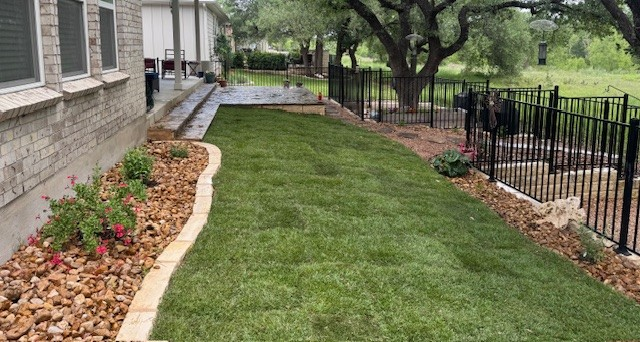 A manicured backyard with fresh green grass, rock beds, and a black fence.