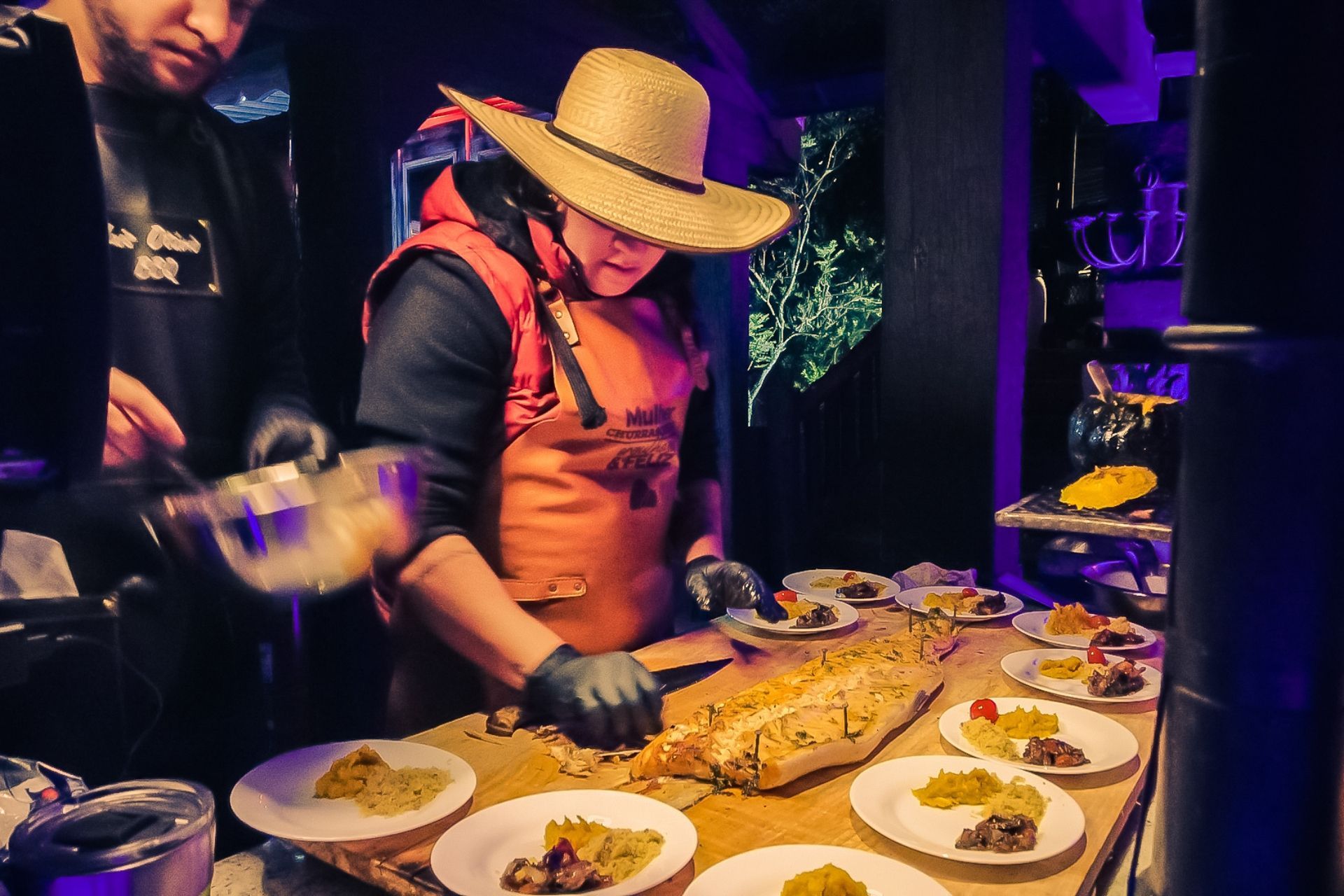 Um homem com chapéu de palha está parado ao lado de uma mesa com pratos de comida.