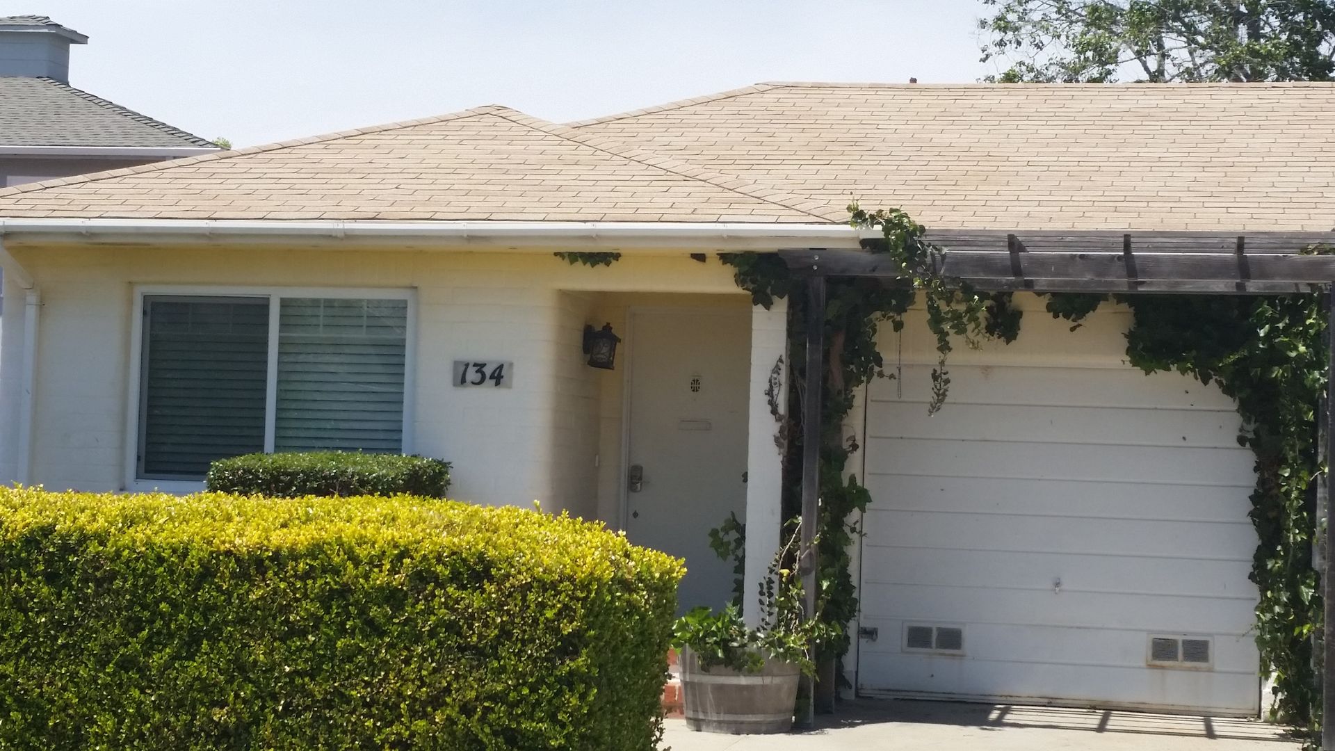 Single-story beige house with white garage door, front hedge, and vine-covered entryway in daylight
