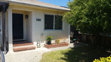 Front of a white house with a porch, dark door, window shutters, and a tree shading the yard.