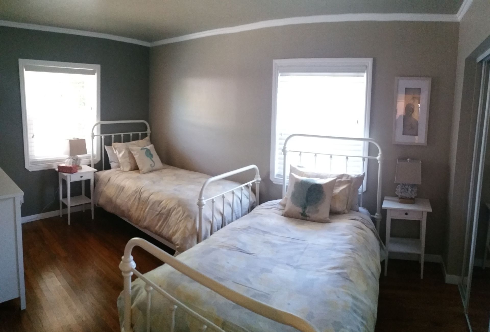 Bright bedroom with two white metal twin beds, gray walls, wood floor, and bedside tables by windows