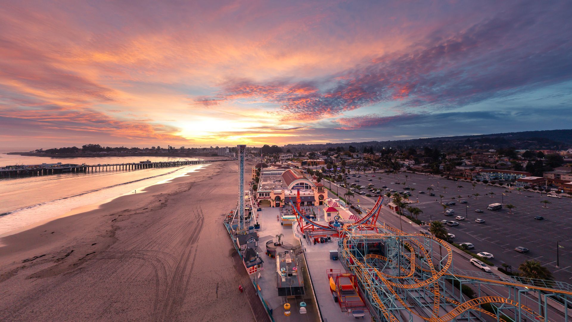 Sunset over a beach boardwalk with colorful lights and the ocean on the left