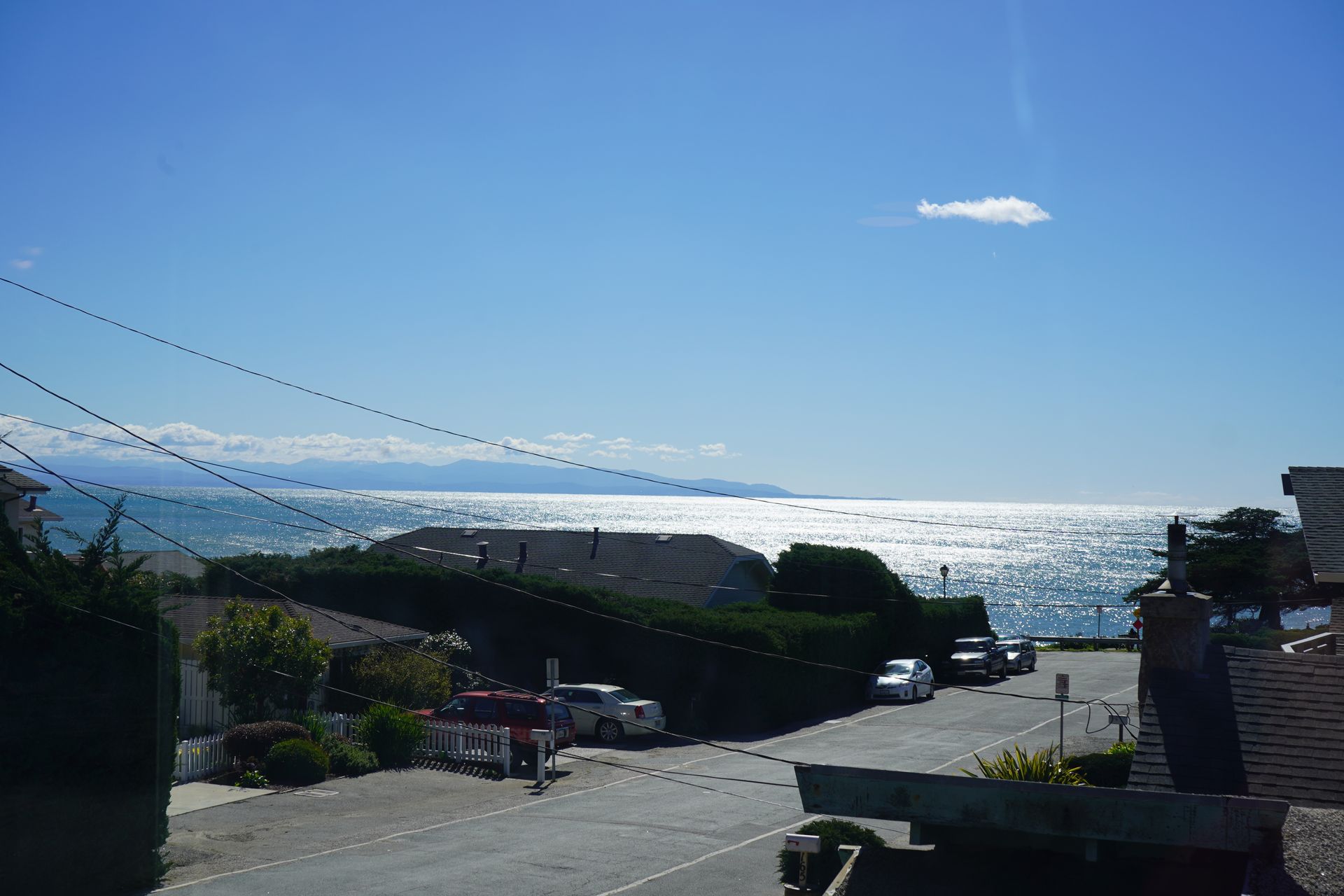 Coastal road overlooking a bright blue ocean under a clear sky, with a few cars and buildings in the foreground
