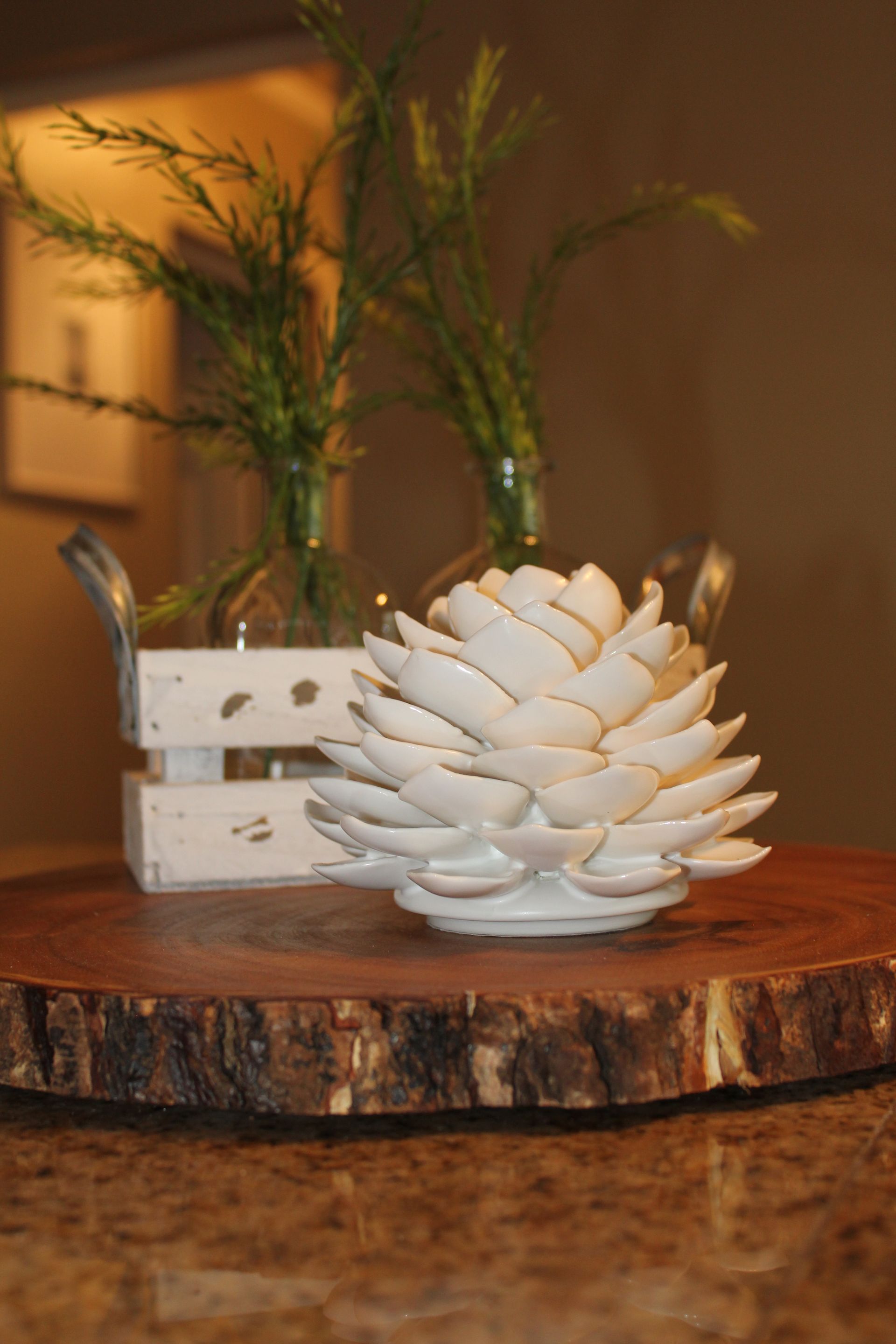 White decorative pinecone candle on a rustic wood slab with a small white crate and greenery in the background