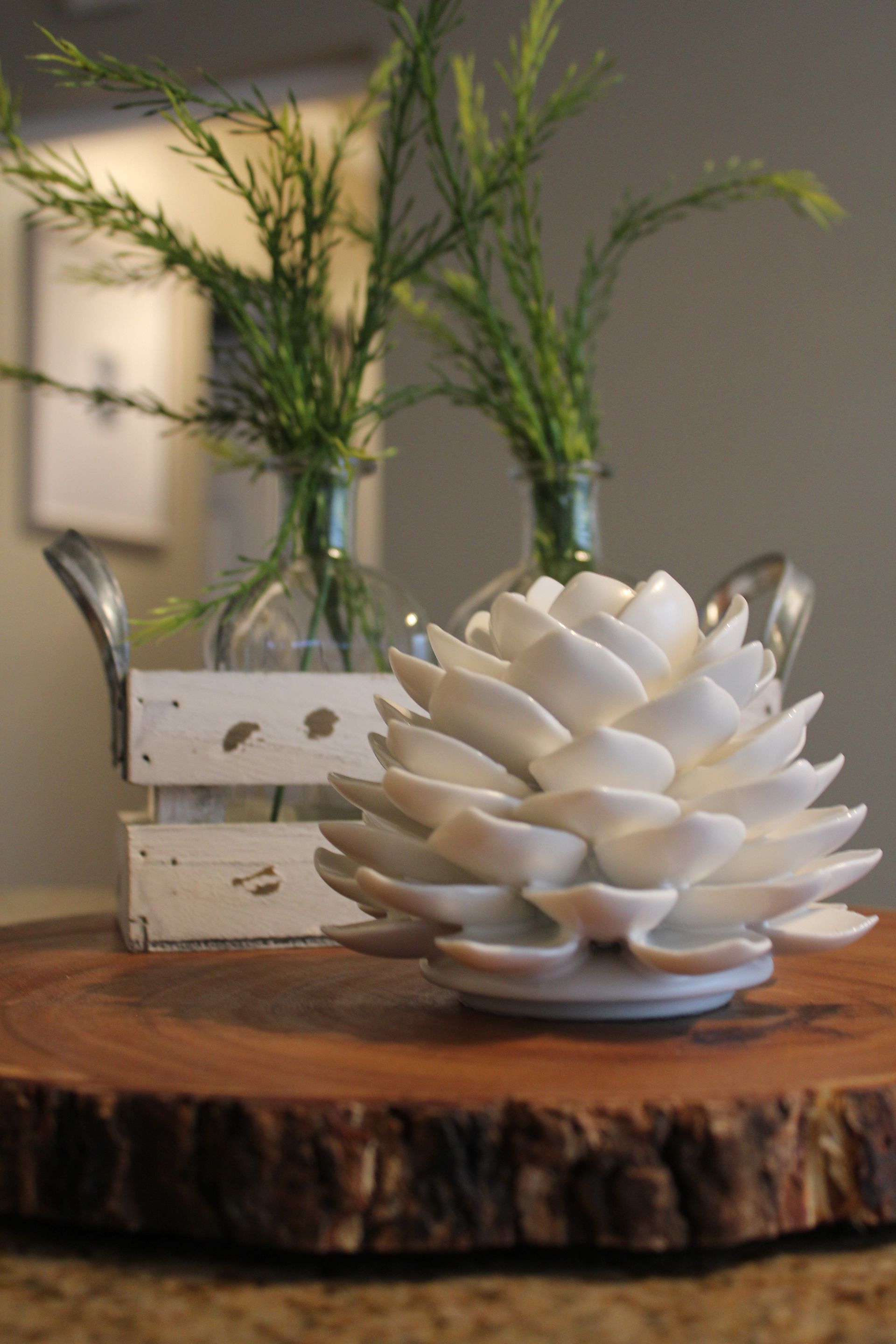 White ceramic flower sculpture on a rustic wood slab, with two small glass vases of greenery in the background
