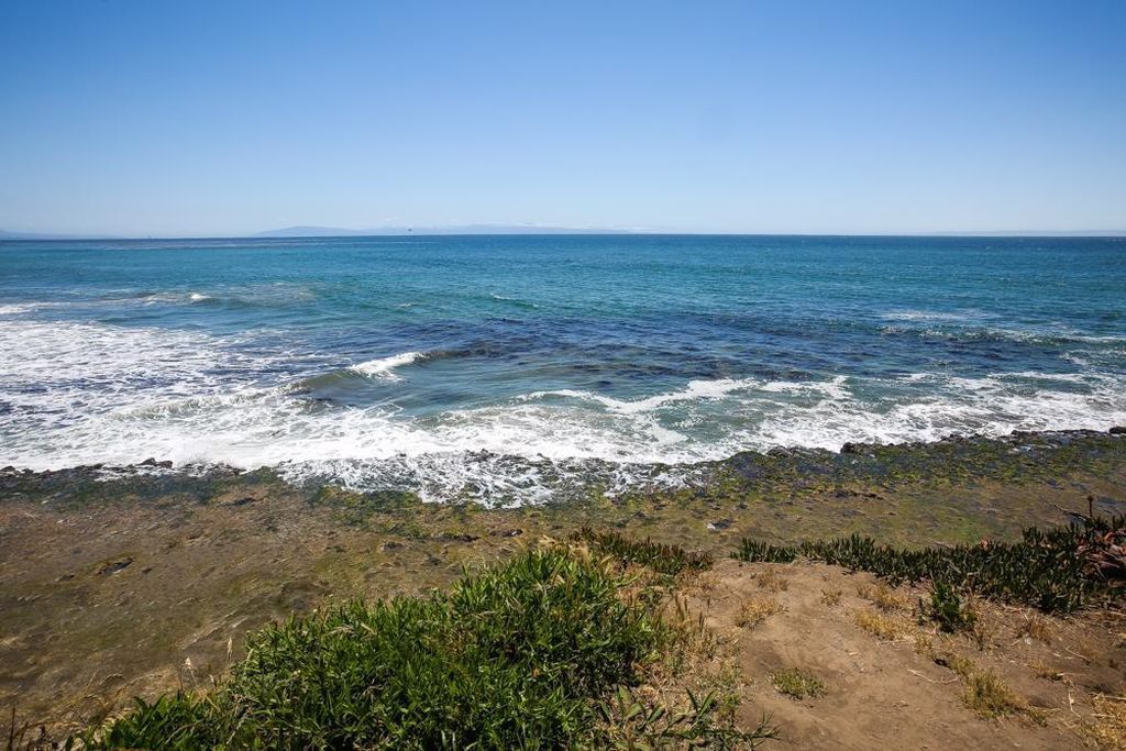 Ocean waves rolling toward a sandy, grassy shoreline under a clear blue sky