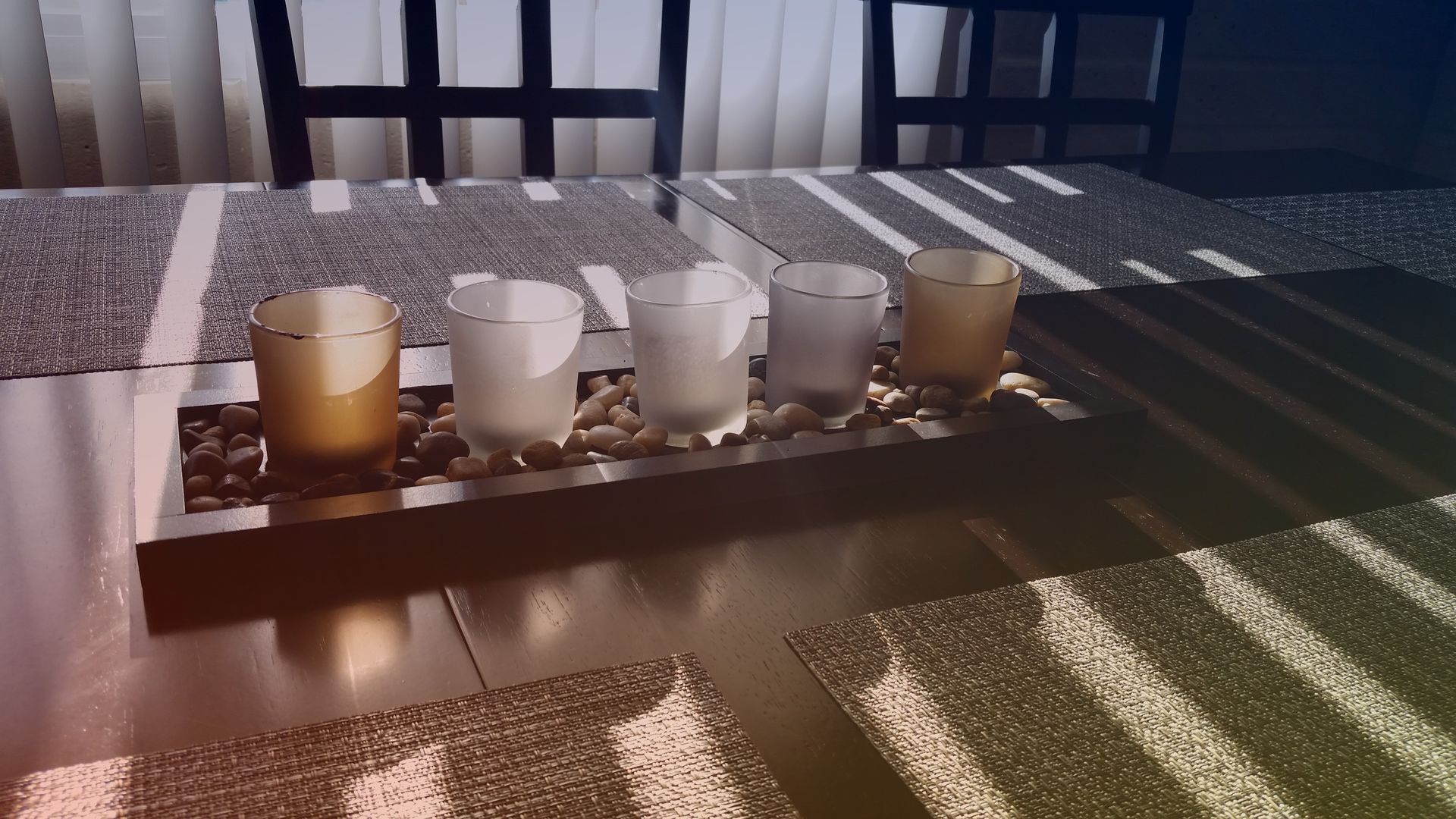 Five small candles in glass holders arranged on a tray atop a dining table with chairs in the background