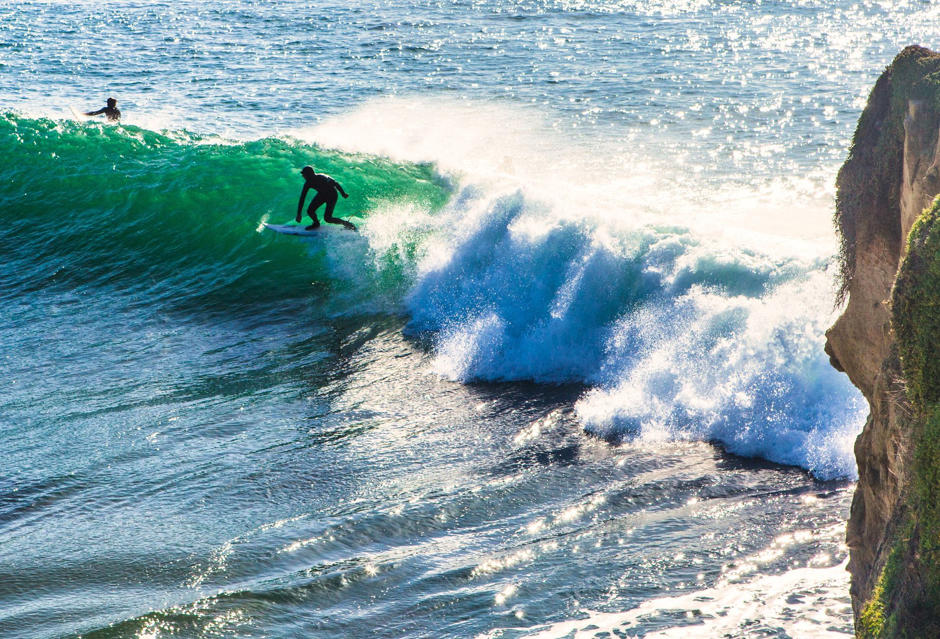 Surfer riding a green wave beside a rocky cliff as white spray crashes