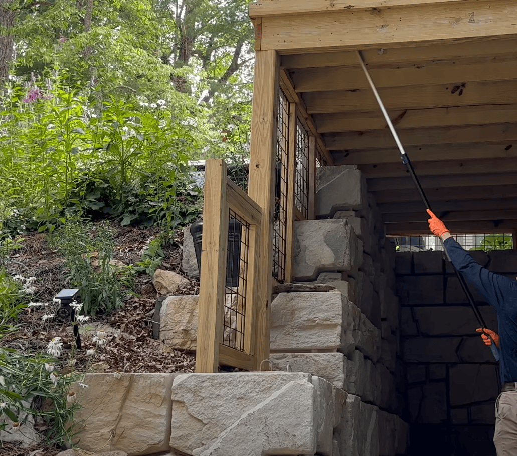 Worker using an extension pole to spray underneath a wooden deck.