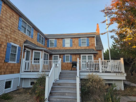 A two-story cedar-shingled house with blue shutters, a wooden deck, and a staircase leading to the entrance at sunset.