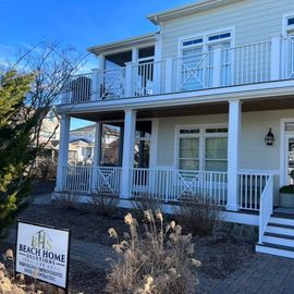A two-story, light green beach house with white balconies and a company sign in the foreground on a sunny day.