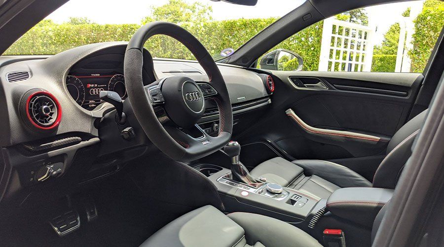 Interior of an Audi RS3. Black and gray leather seats, steering wheel, and dashboard. Red accents.
