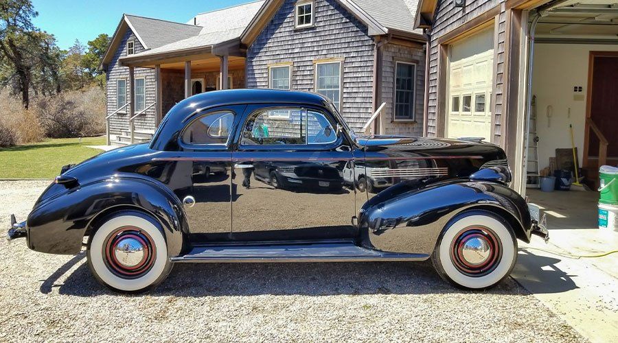 Black vintage car with white-wall tires parked in front of a wooden house and garage.