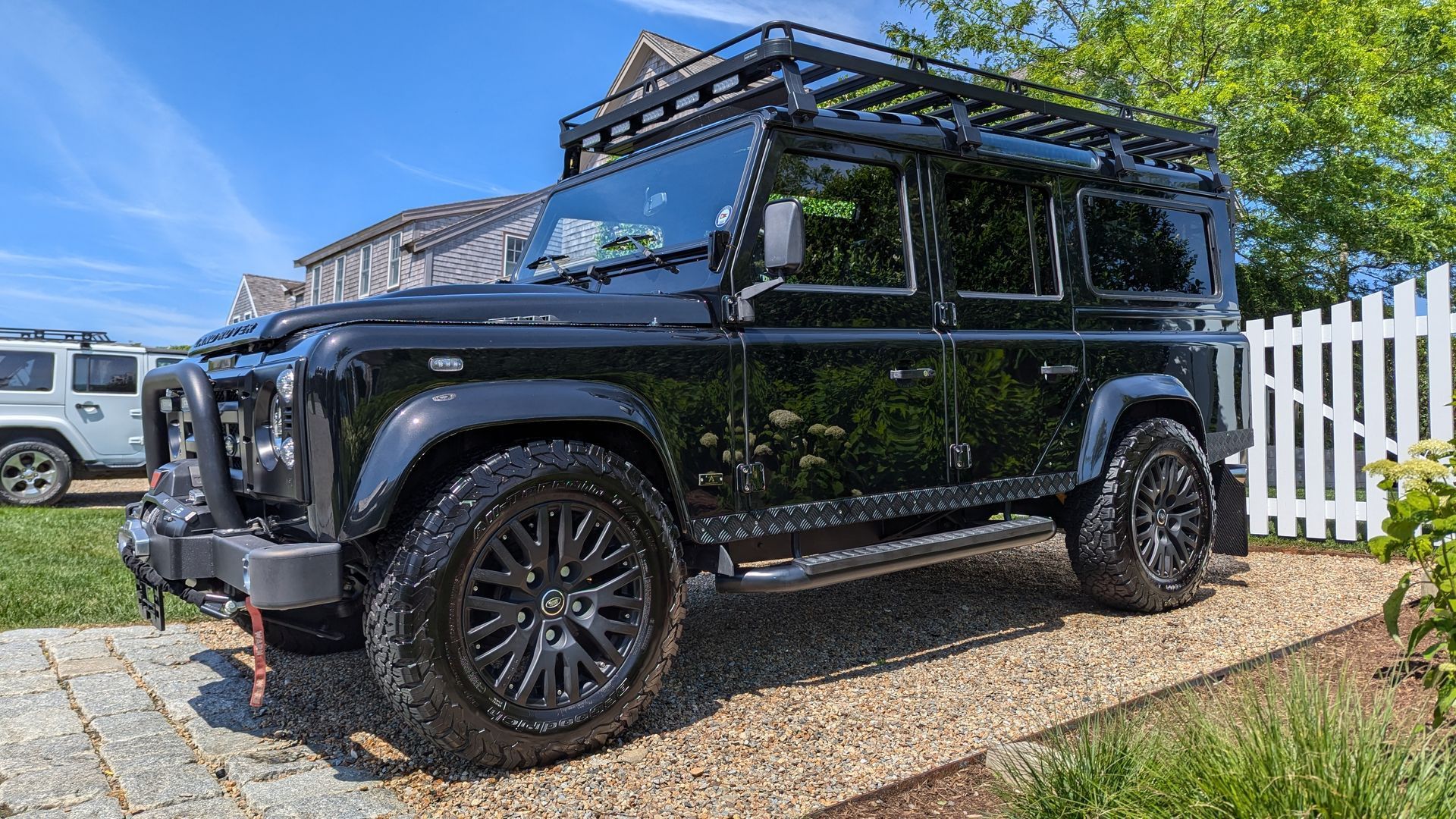 A black land rover defender is parked in a driveway next to a white picket fence.
