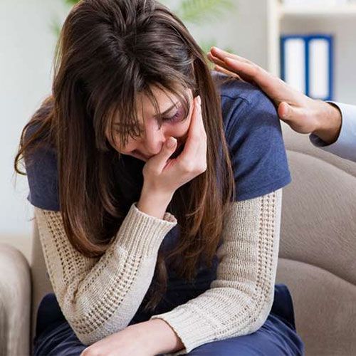 A woman is sitting on a couch with her head in her hands and a man is comforting her.