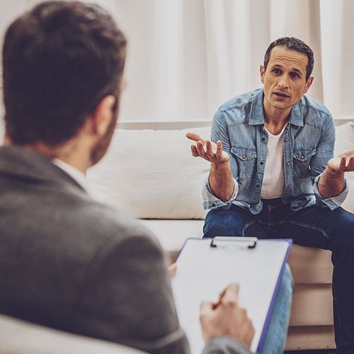 A man is sitting on a couch talking to a man holding a clipboard.