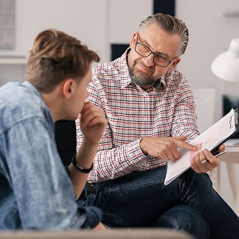 A man is sitting on a couch talking to another man while holding a clipboard.