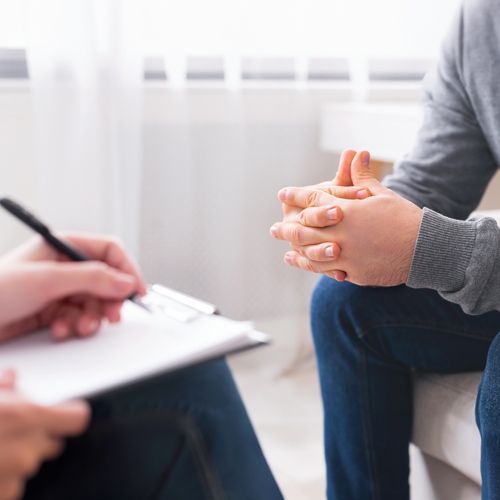 A man is sitting on a couch with his hands folded while a woman writes on a clipboard.