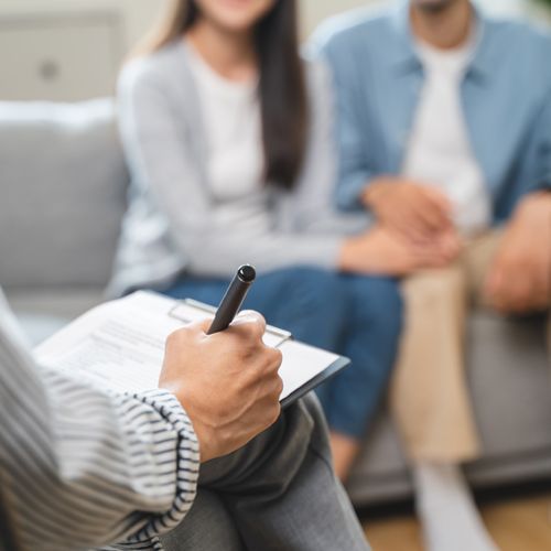 A woman is writing on a clipboard while a man and woman sit on a couch.