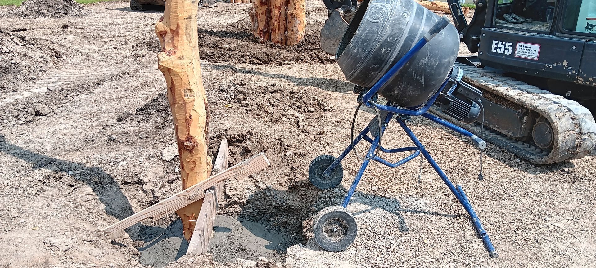 A concrete mixer is sitting in the dirt next to a tree.