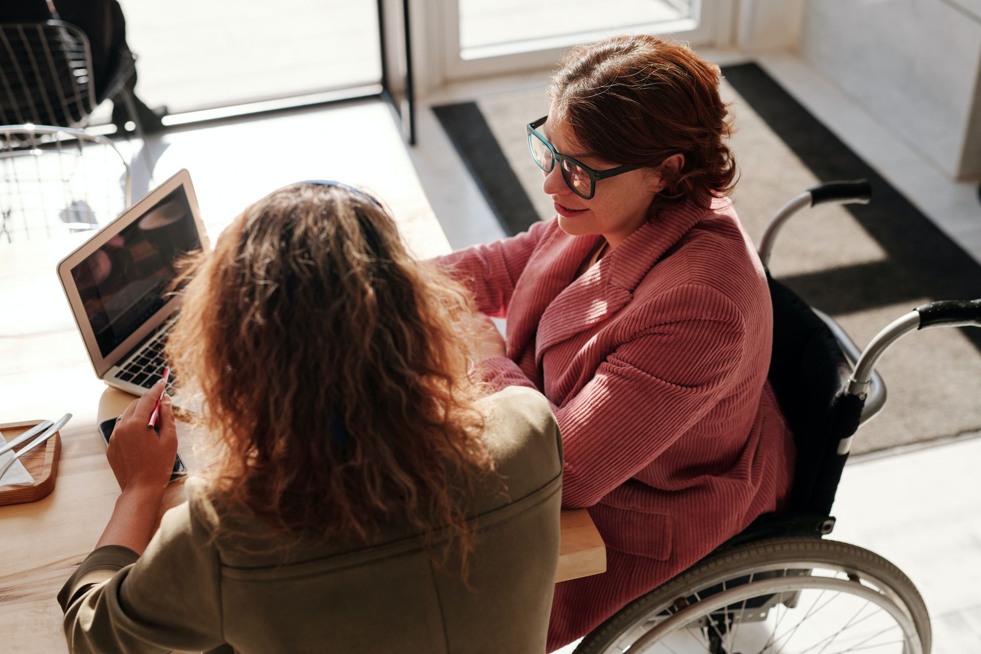 Two people sit at a table working on a laptop, one using a wheelchair, in a bright, modern office space.
