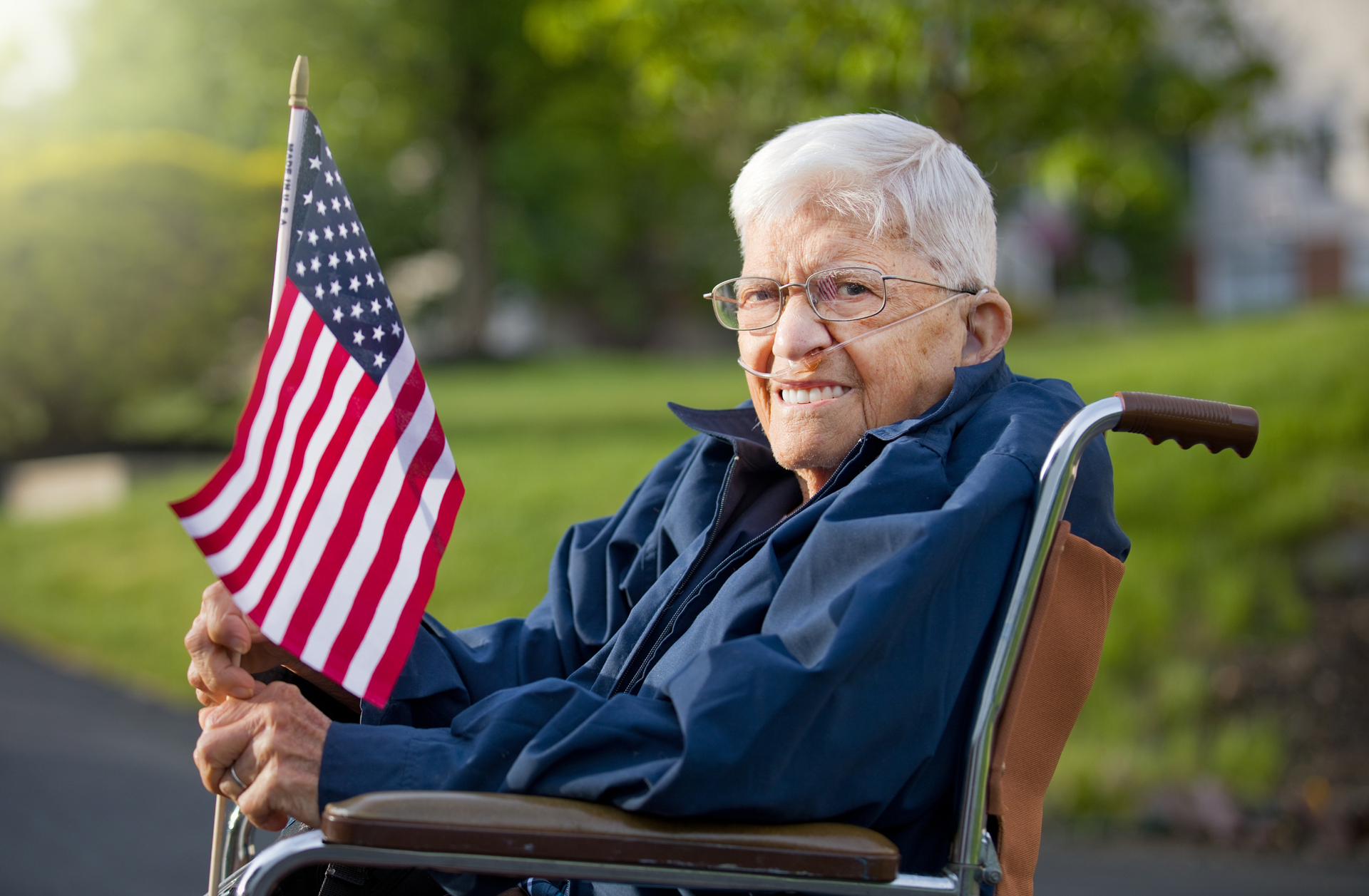 A person in a wheelchair wearing a blue jacket holds an American flag while sitting outside in a grassy area.