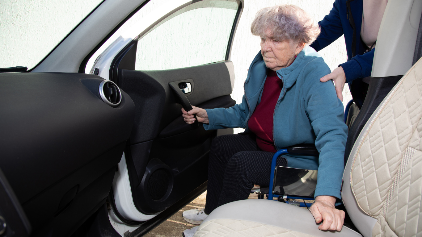 A person in a blue jacket is assisted in transferring from a wheelchair to the front seat of a car.