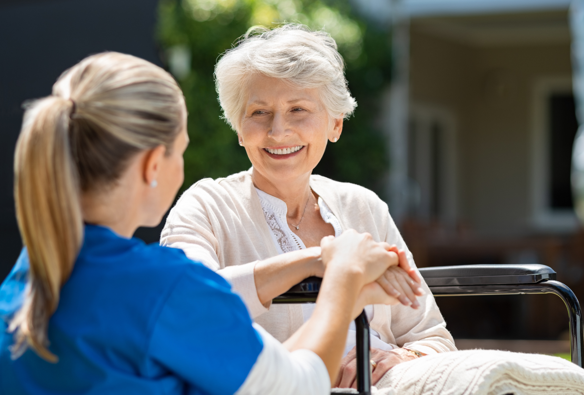 A healthcare provider in a blue uniform holds the hand of a smiling individual seated in a wheelchair outdoors.