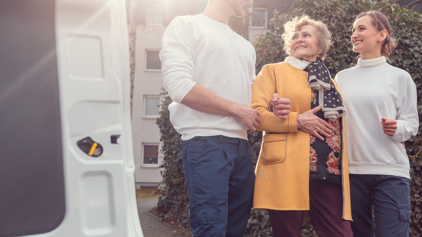 A person in a yellow coat walks arm-in-arm with two others outdoors near a white vehicle.