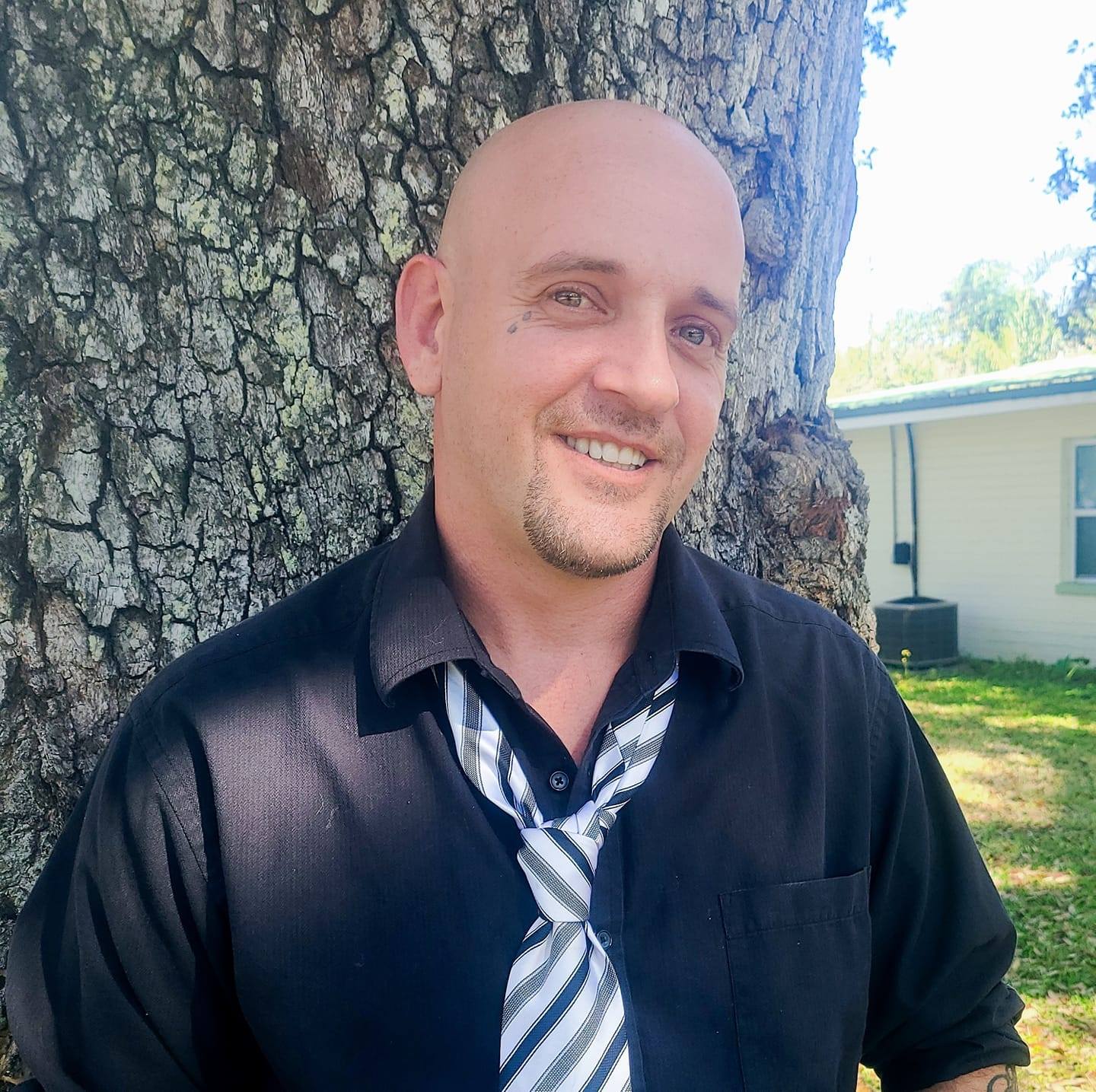 Jake the co owner smiles while leaning against a tree. He wears a black shirt and patterned tie.