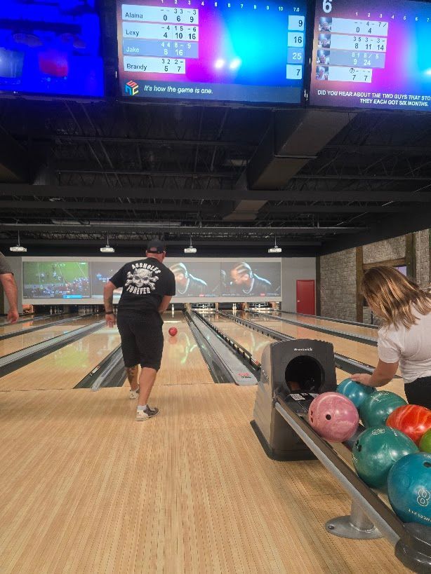 People bowling at an alley; man bowling, woman getting balls; lane with scoreboards overhead.