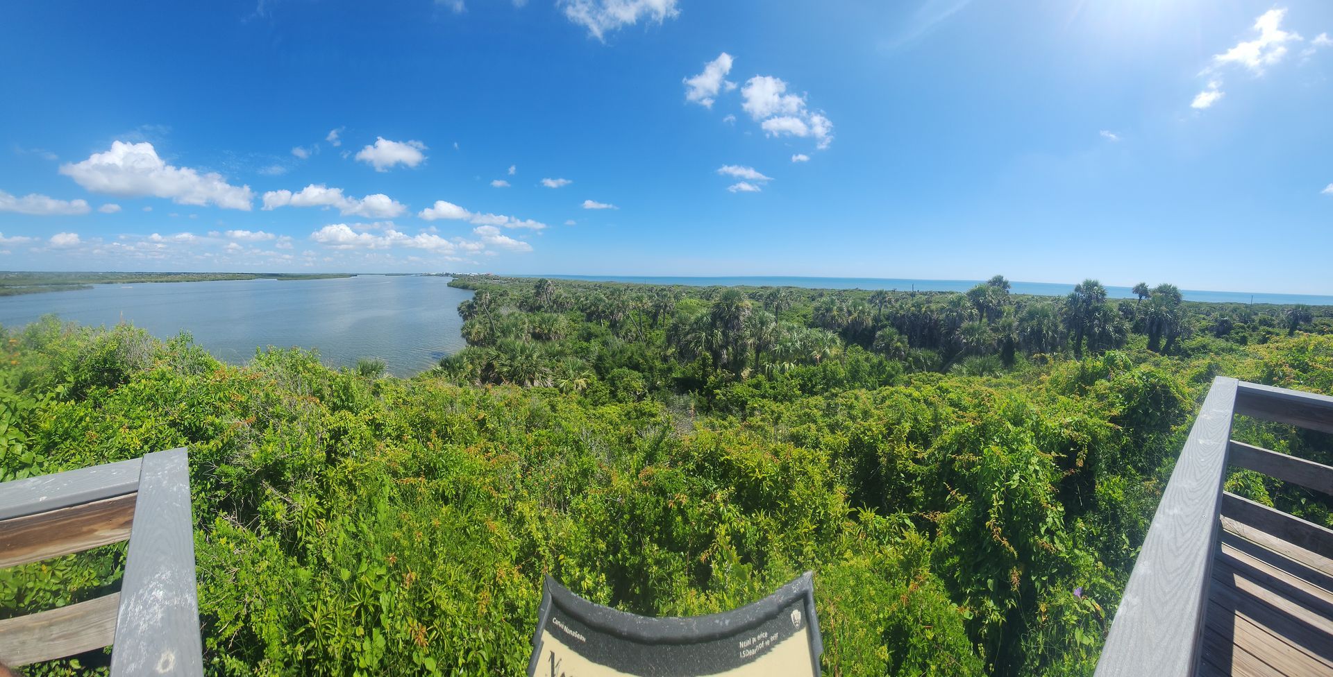 View from an observation deck overlooking a blue lake, lush green trees, and blue sky with clouds.