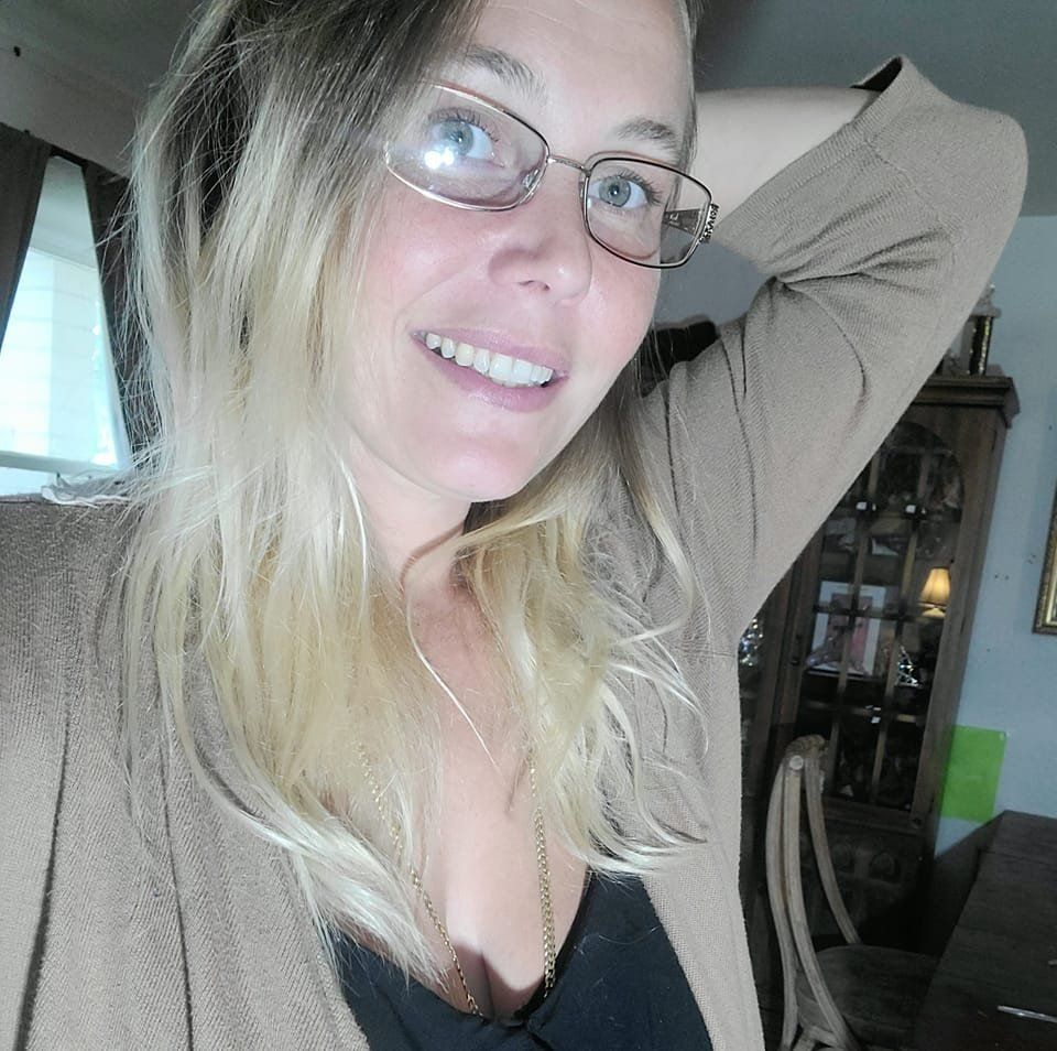 Woman with blonde hair and glasses smiling, arm raised, wearing a tan cardigan and black top.