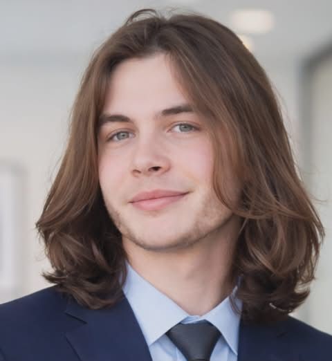 Young man with long brown hair, wearing a black hoodie, blue pants, and a necklace, sitting on a rocky surface.