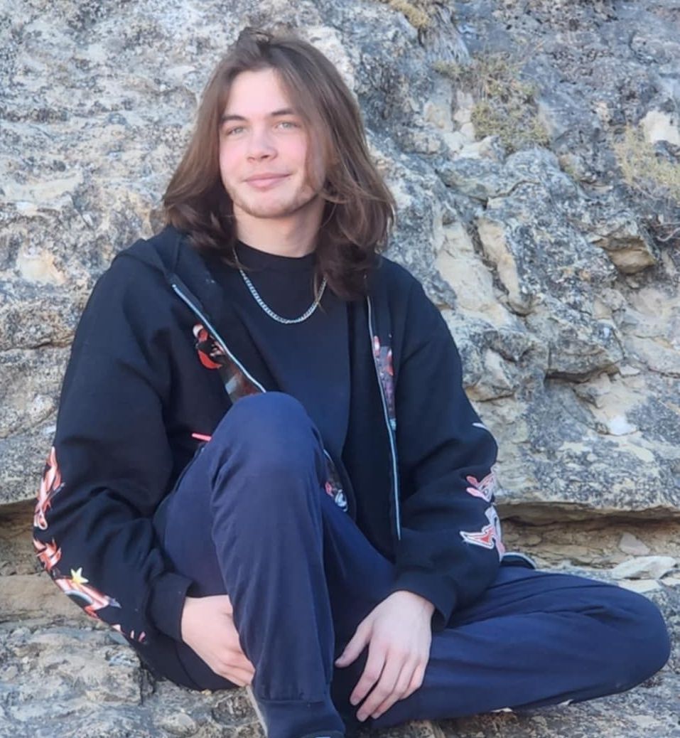 Young man with long brown hair, wearing a black hoodie, blue pants, and a necklace, sitting on a rocky surface.