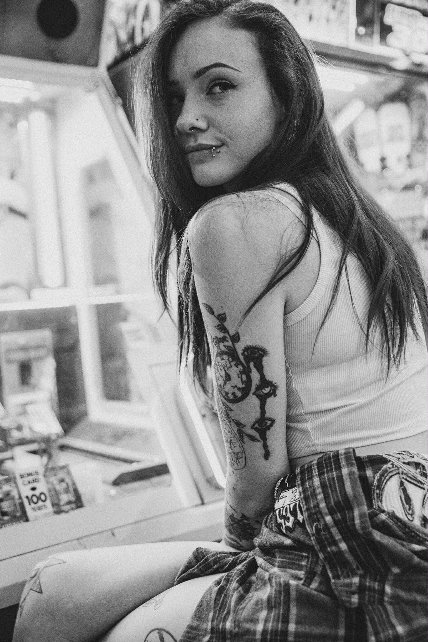 A young woman with long dark hair, tattoos, and a white tank top smiles at the camera. She sits inside a store.