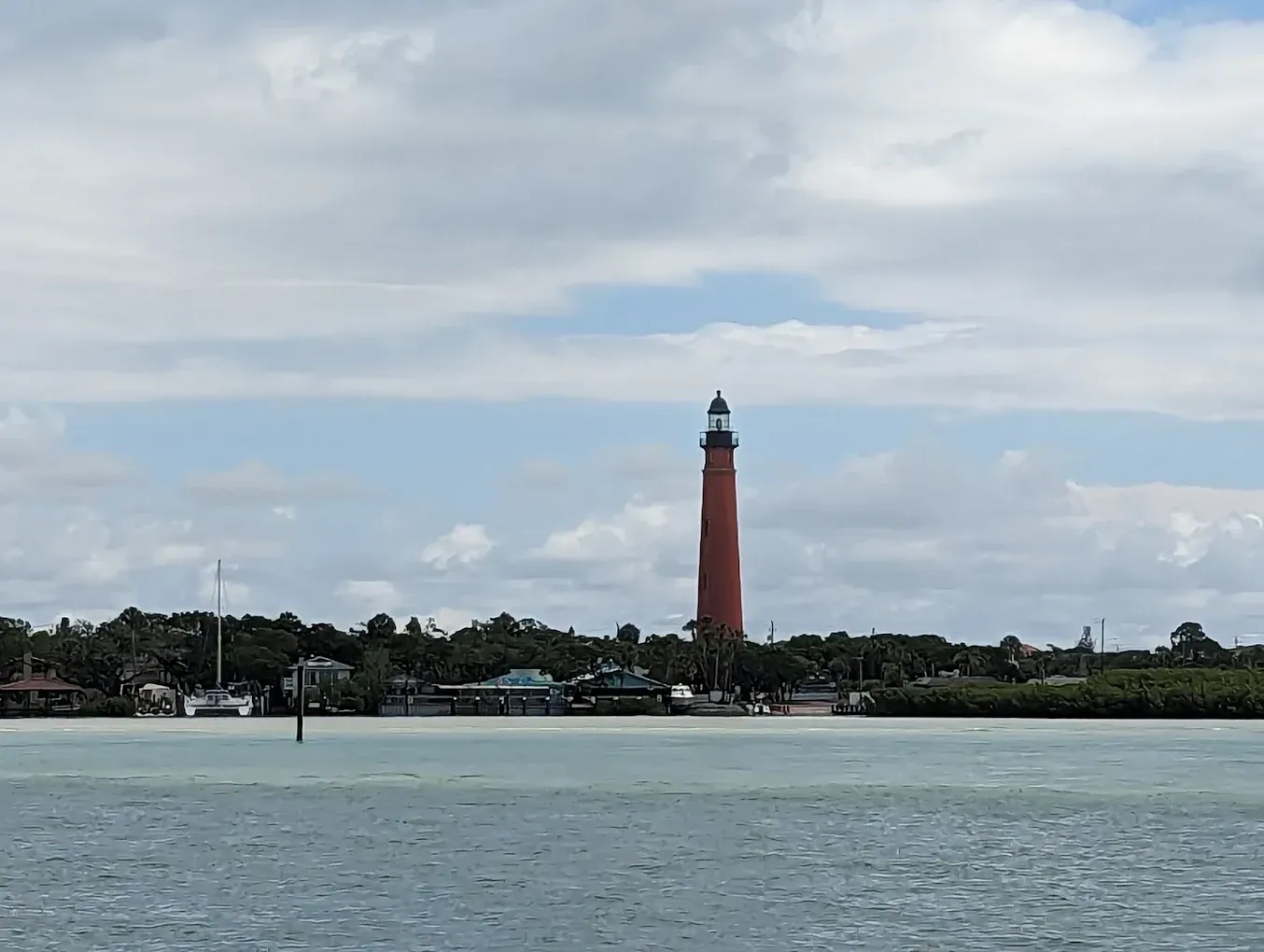 Tall, red lighthouse stands against a cloudy sky, overlooking a coastline and water.