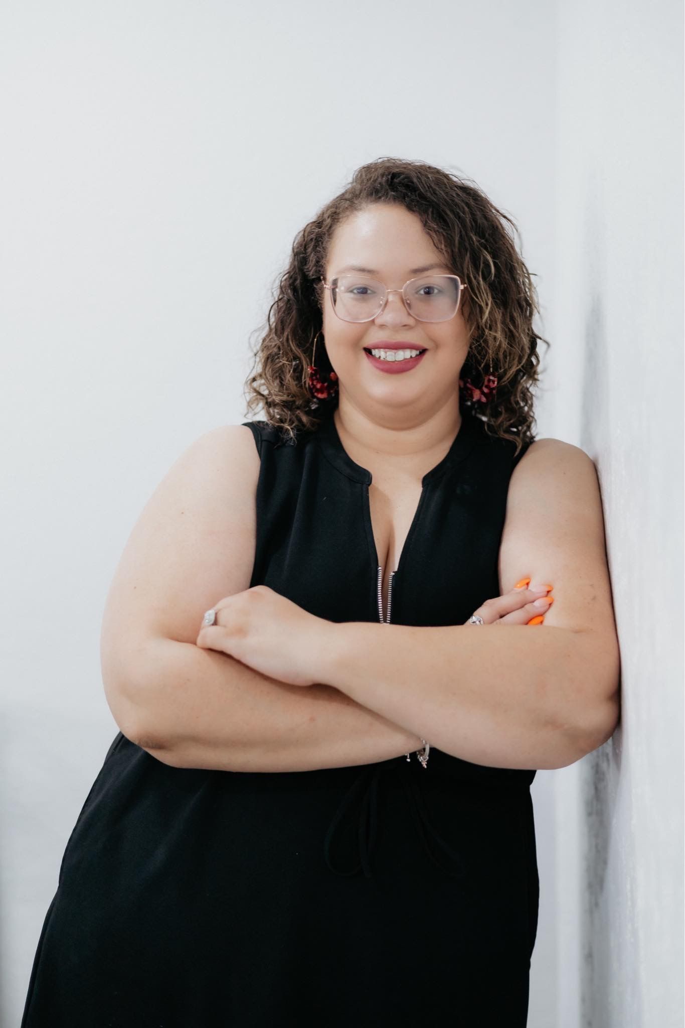 A woman in a black dress and glasses is leaning against a wall with her arms crossed.