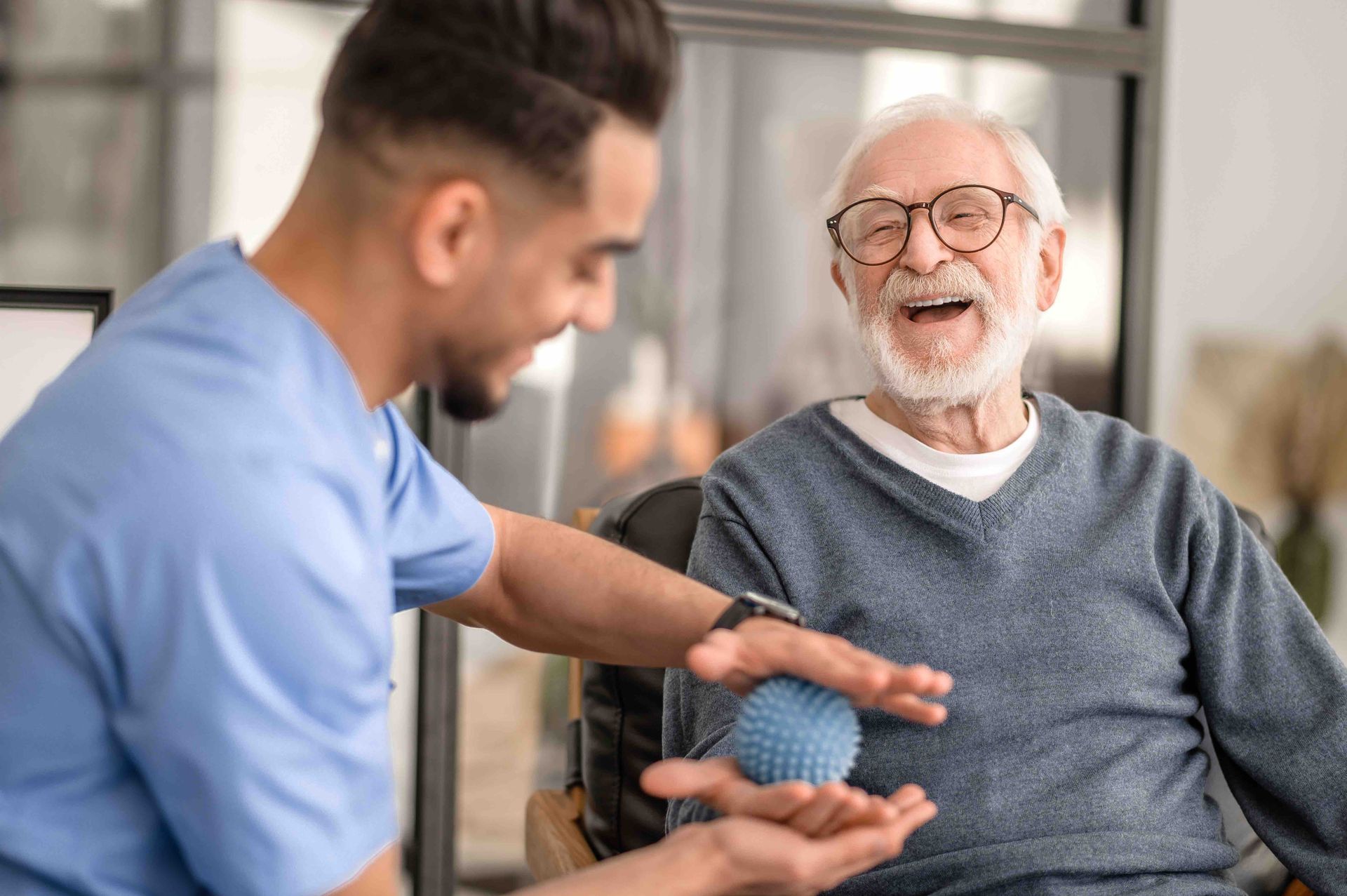 A medical professional in blue scrubs helps an older man perform hand exercises with a blue textured therapy ball.