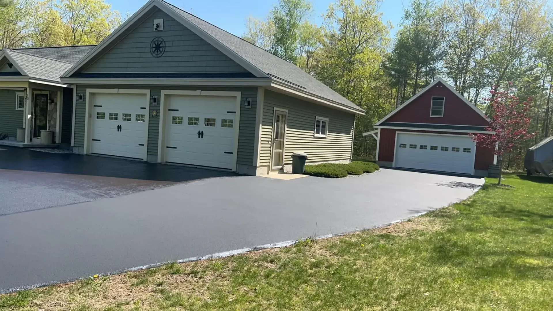 A gray house with a two-car garage and asphalt driveway, adjacent to a red garage, under a blue sky.